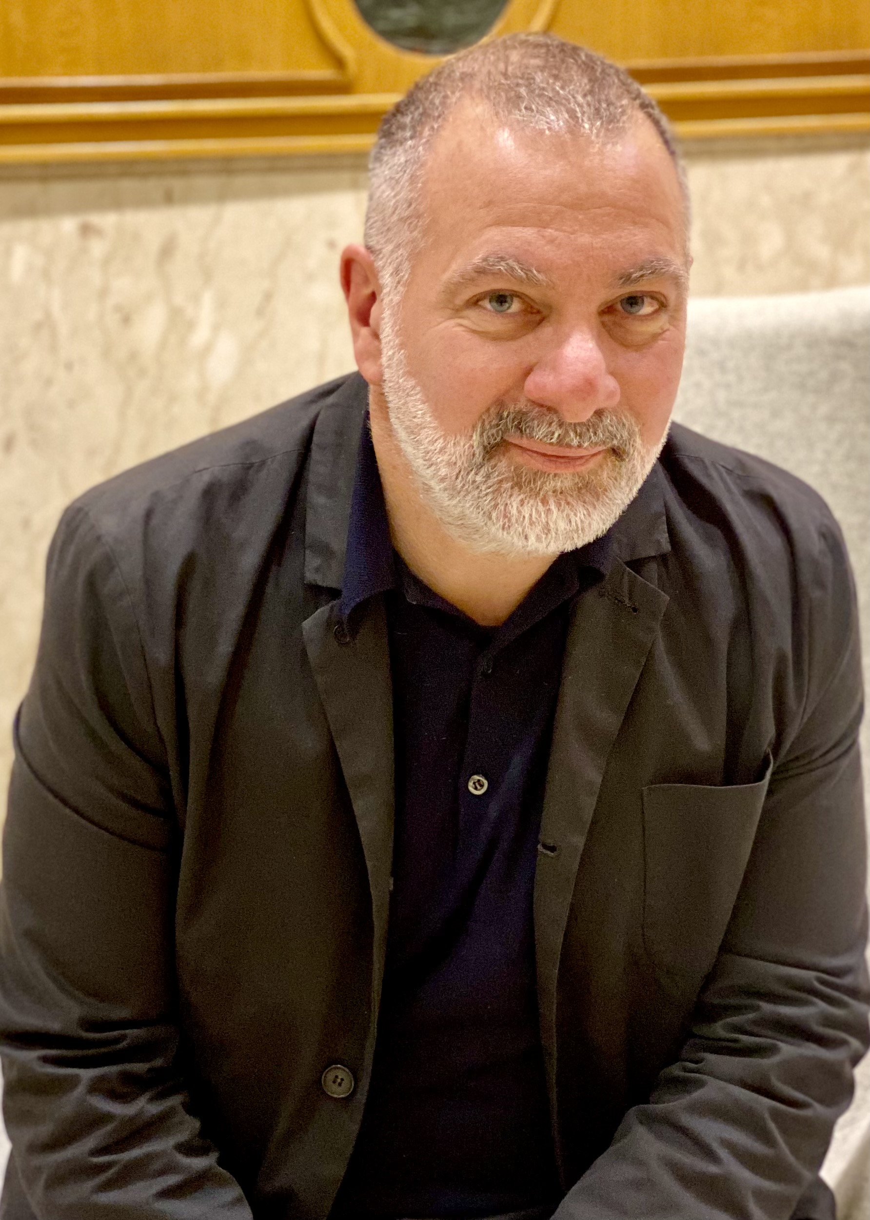Portrait photo of a man with short gray hair and beard, wearing a black jacket, sitting indoors.