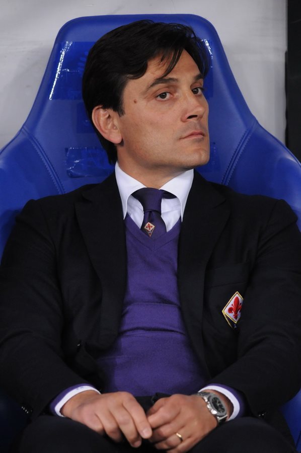 Vincenzo Montella sitting in a blue chair, wearing a black jacket with a purple sweater and tie featuring the ACF Fiorentina club logo.