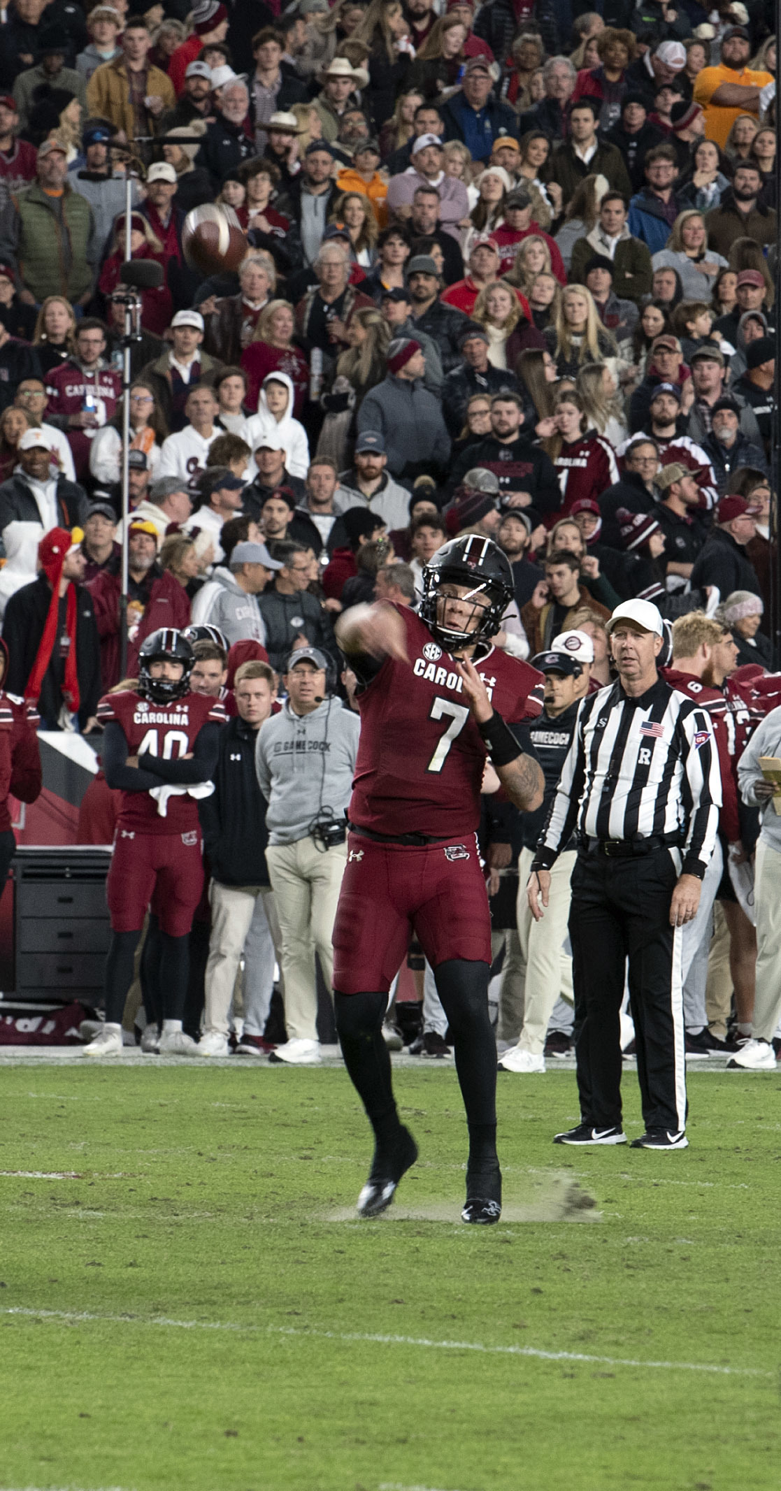 A football quarterback in a dark garnet South Carolina uniform throws a football during a game with a crowd in the background.