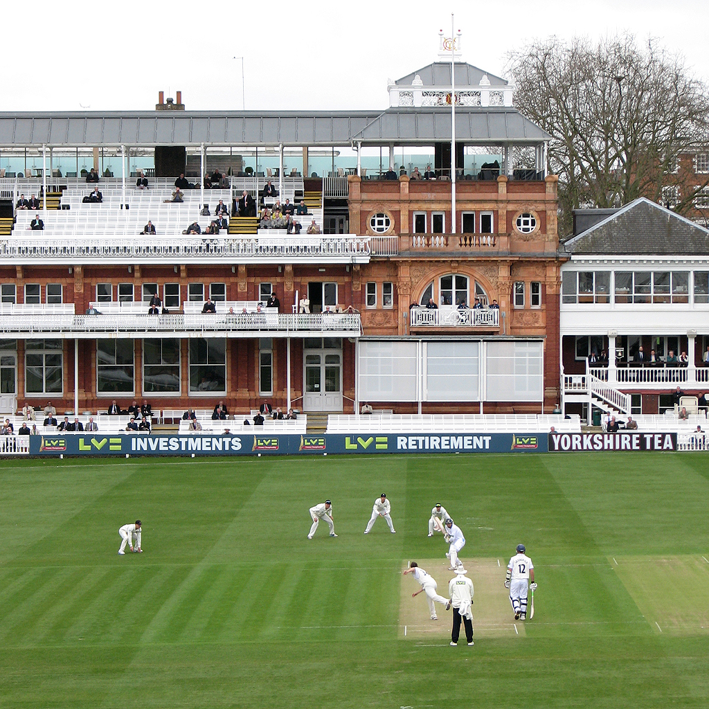 Cricket match at Lord's with bowler bowling to batsman and fielders positioned on the field in front of a pavilion.