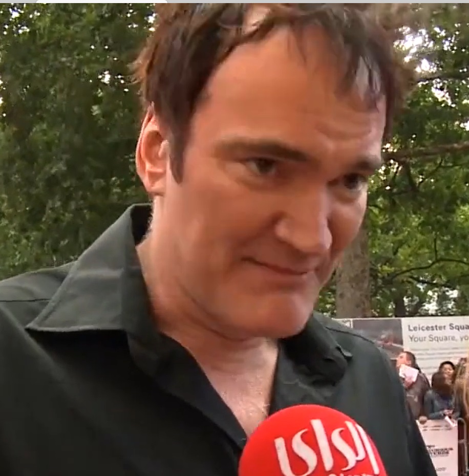 Close-up photograph of a man in a black shirt speaking into a red microphone outdoors with trees in the background.