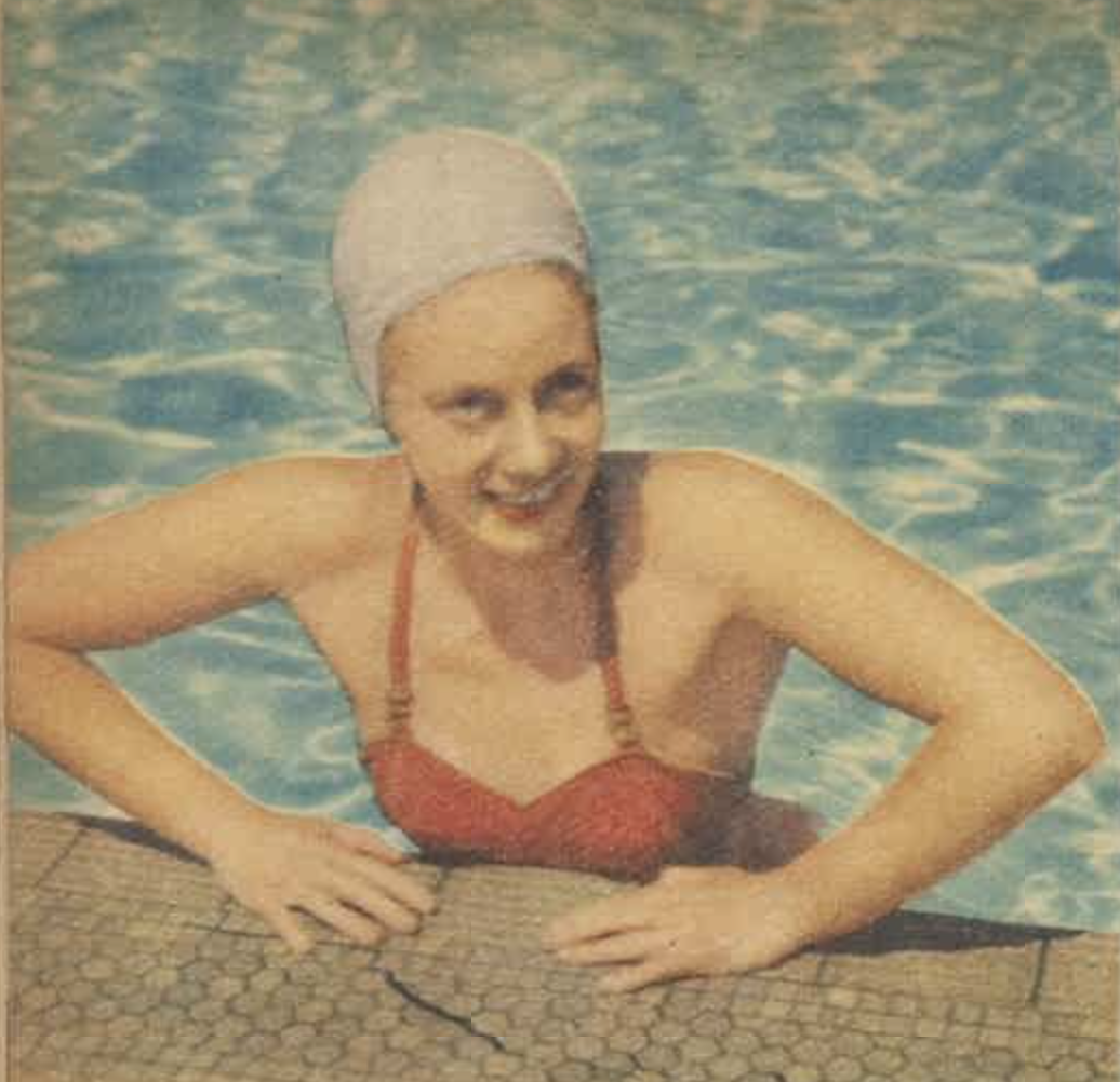 A woman in a red swimsuit and white swim cap emerging from a swimming pool, smiling at the camera.