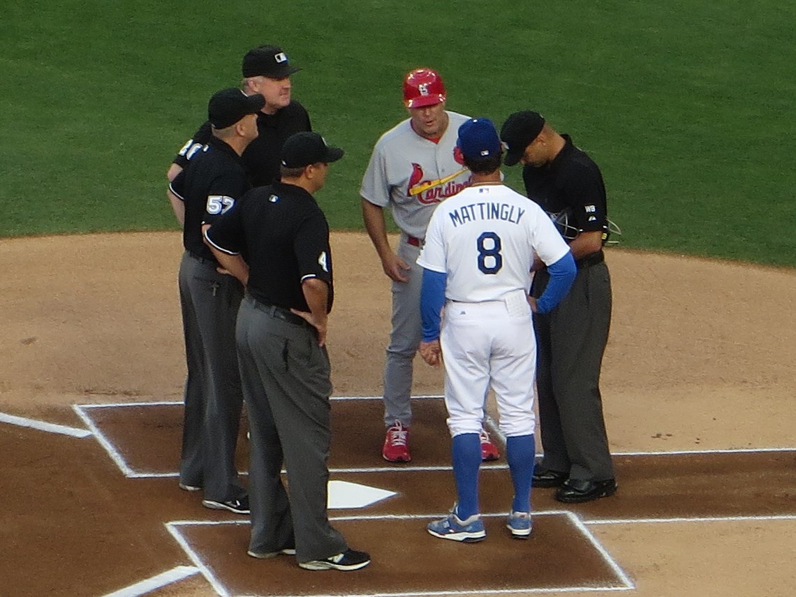 Don Mattingly in a Dodgers uniform standing on a baseball field with other baseball umpires and a player in a Cardinals uniform.