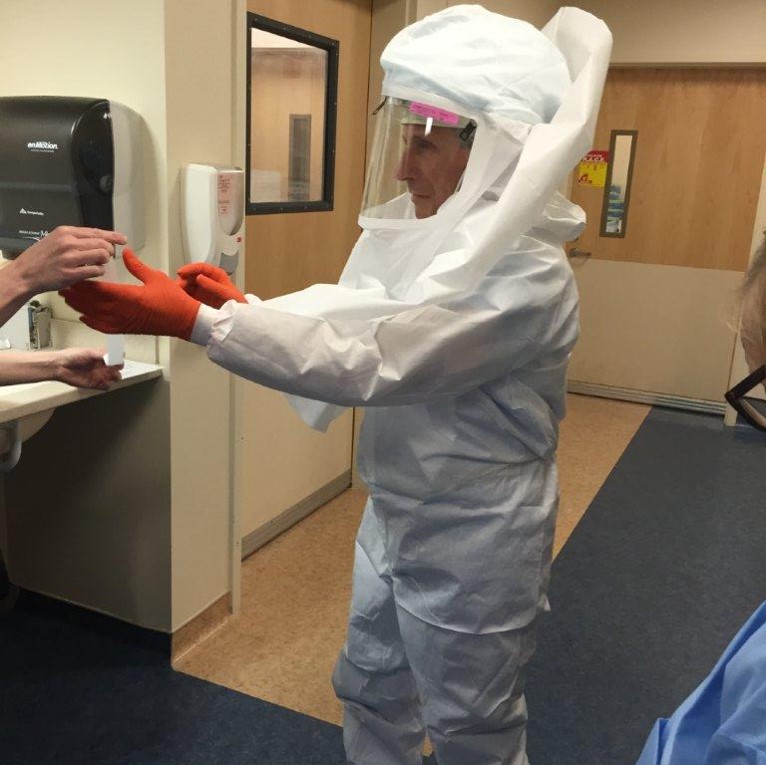 Person wearing white full-body personal protective equipment with a clear face shield and orange gloves inside a medical facility.