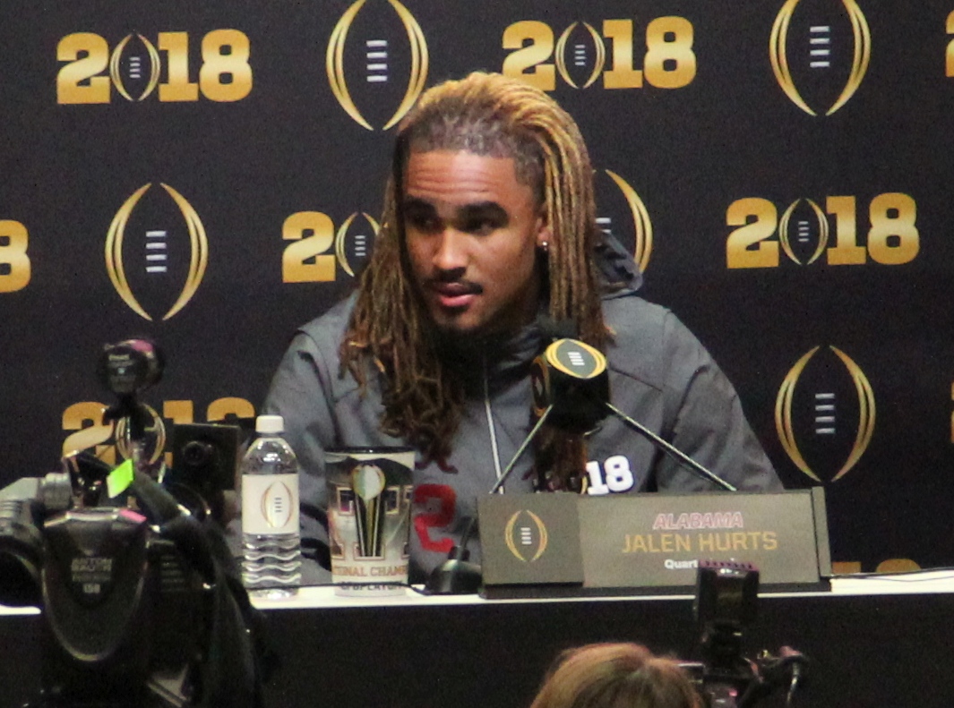 Jalen Hurts at a 2018 media day press conference with microphones and a nameplate in front of him
