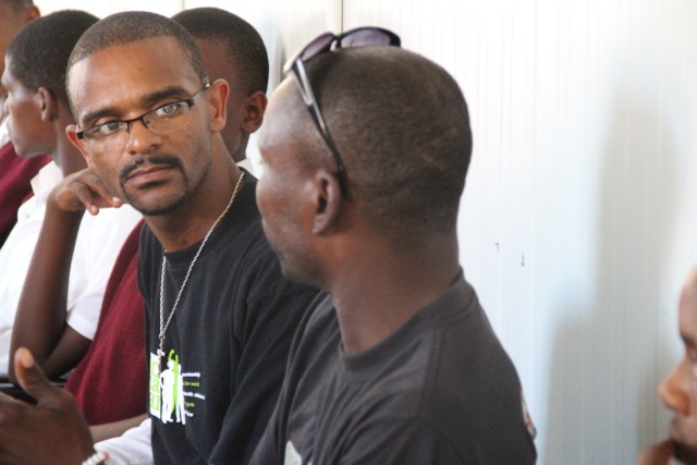 Several men sitting in a row, engaged in conversation, one wearing glasses and a black shirt with a visible campaign logo.