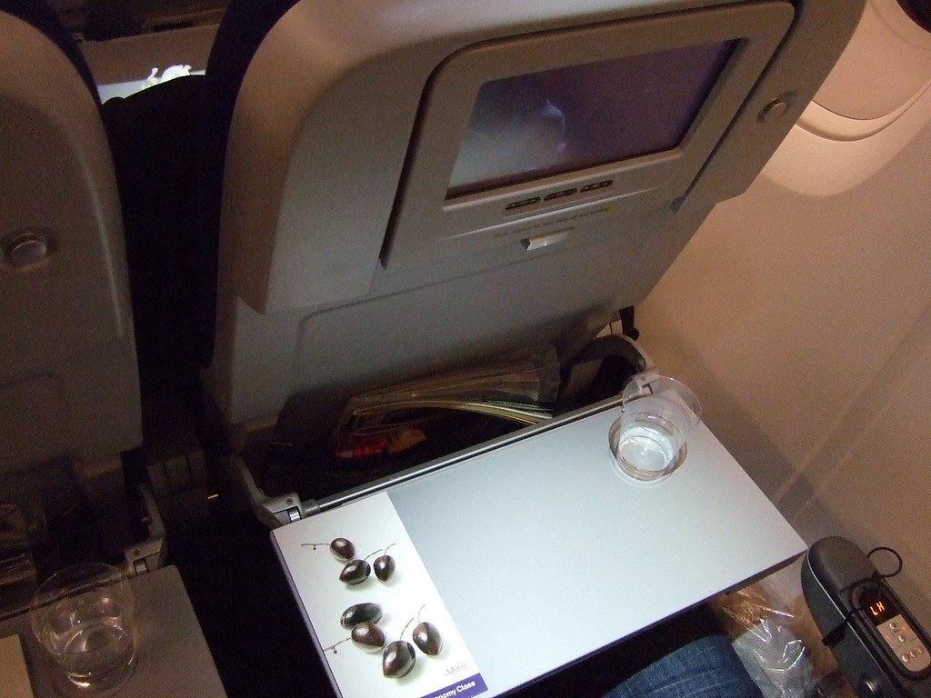 Economy class airplane seat back with in-flight entertainment screen, folding tray table with water and cherries, and part of passenger's lap and armrest visible.