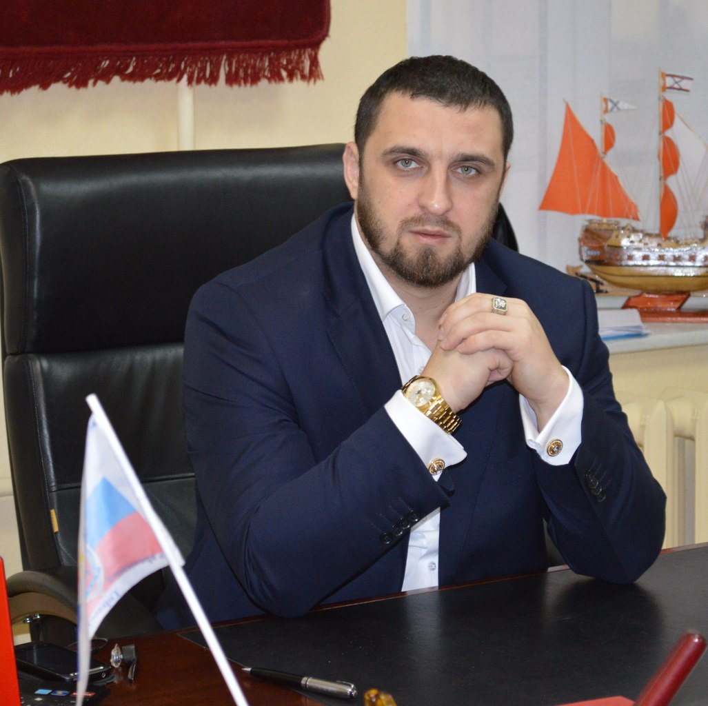 A man with short dark hair and beard wearing a dark suit and white shirt, sitting at a desk with hands clasped, a small flag and model ships in the background.