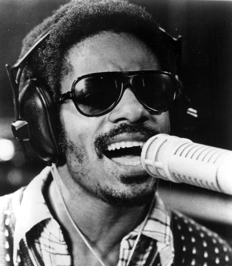 Black and white close-up photograph of Stevie Wonder wearing sunglasses and headphones, singing into a microphone.
