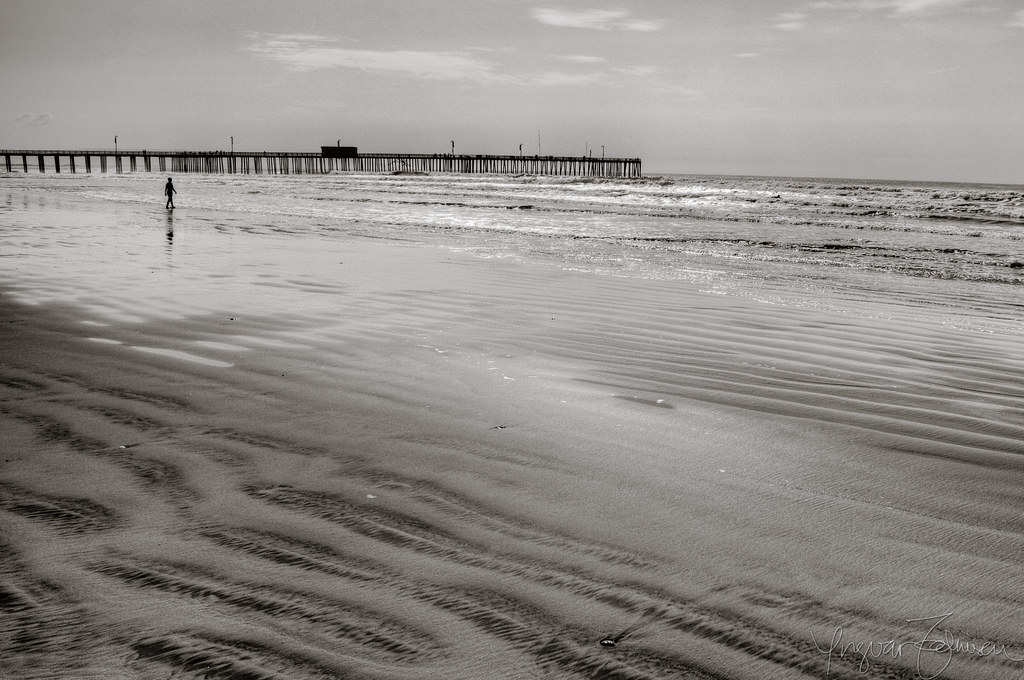 Black and white photo of a sandy beach with water ripples and a long pier extending into the ocean, with one person walking near the water.