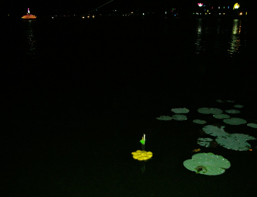 An edible yellow Krathong floating on dark water at night near lily pads with distant lights reflected on the water.