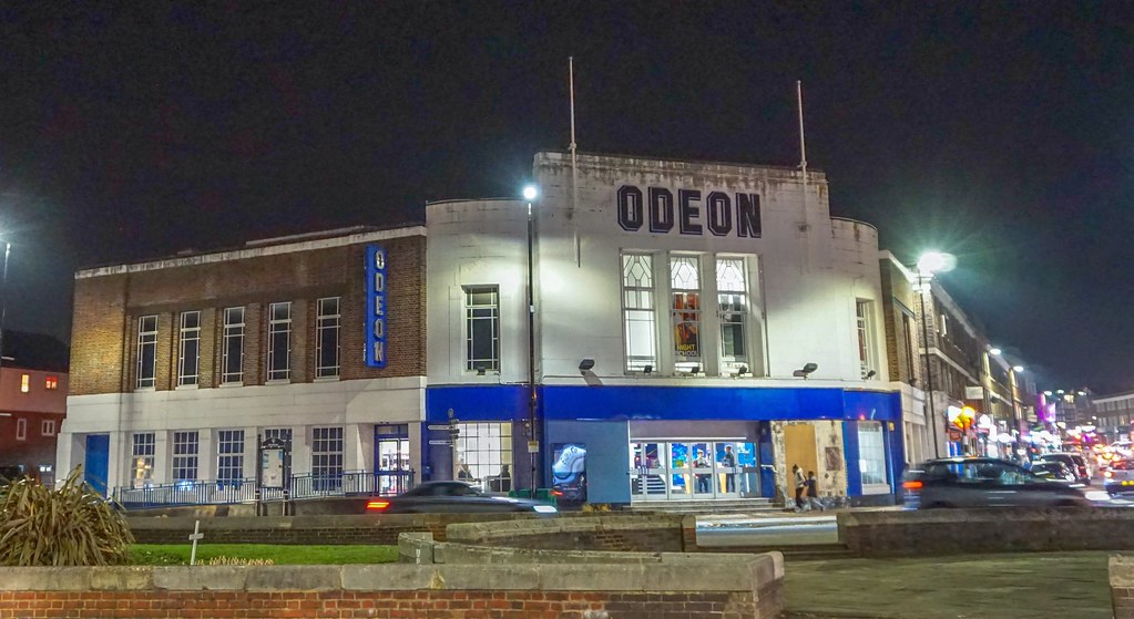 The Beckenham Odeon cinema building at night with lit 'ODEON' signs and cars passing by.
