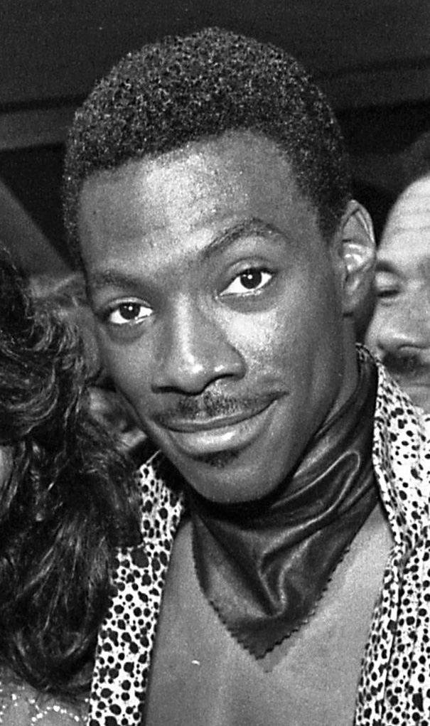 Black and white photograph of Eddie Murphy in 1984, close-up showing his face and upper shoulders, wearing a patterned shirt and scarf.