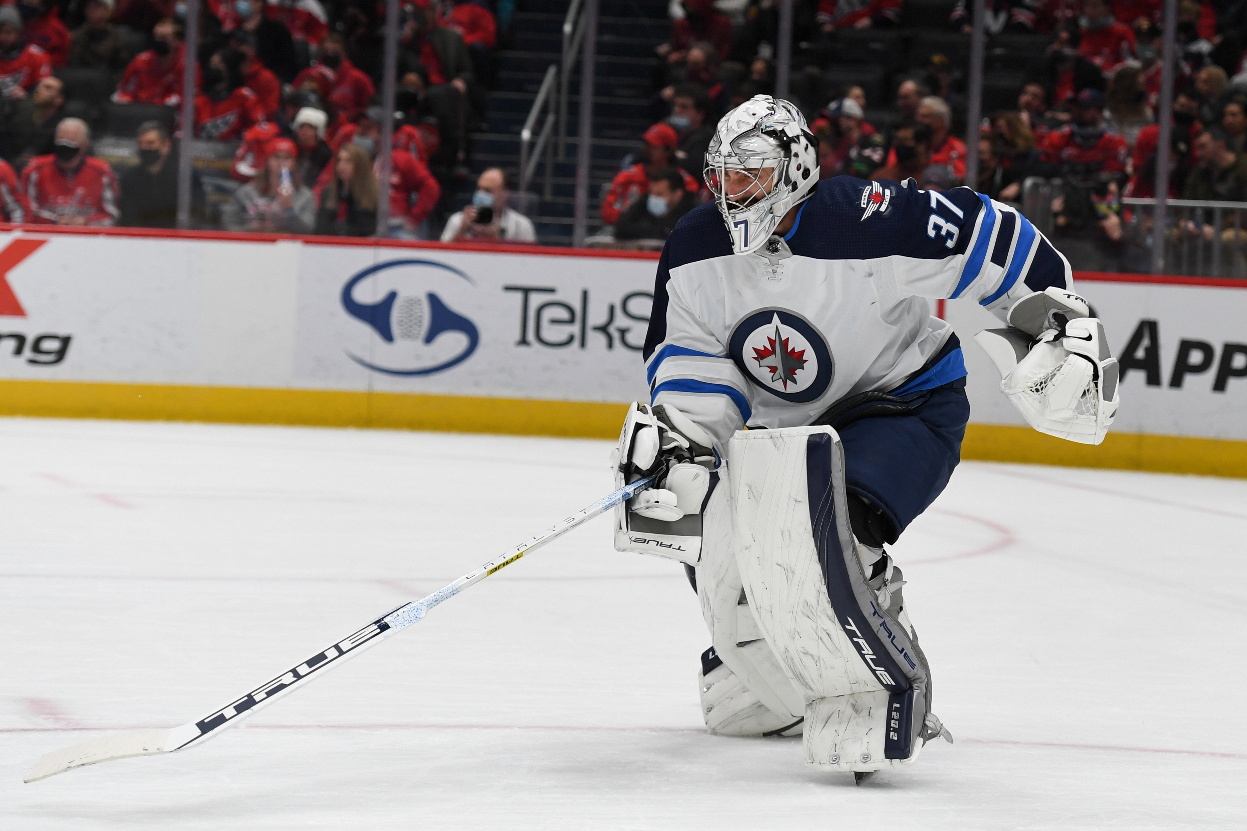 Ice hockey goalie in white and blue Winnipeg Jets jersey with number 37, on ice during game, holding goalie stick and wearing goalie mask.