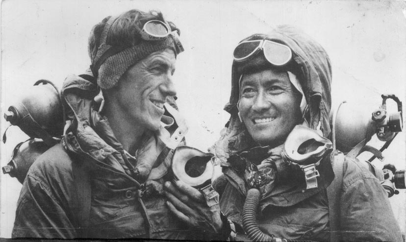 Black and white photograph of two climbers in mountaineering gear, smiling and looking at each other, with oxygen masks around their necks.