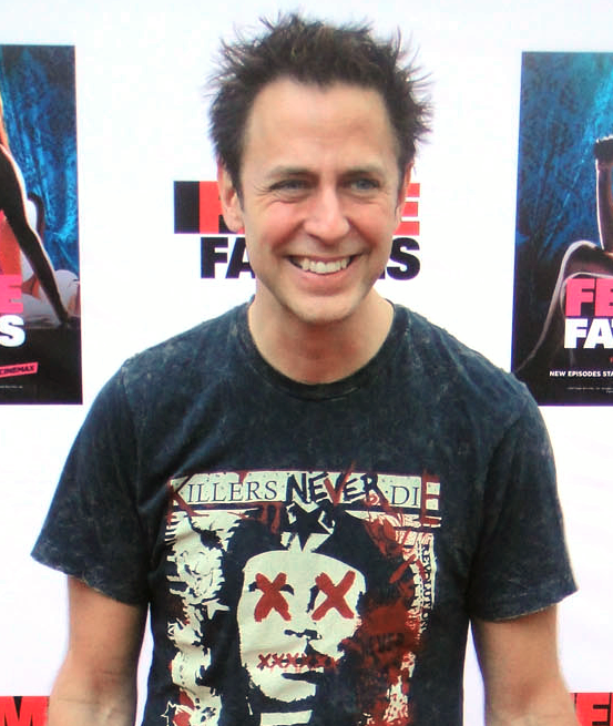 Smiling man with spiked dark hair wearing a graphic T-shirt, standing against a backdrop with text