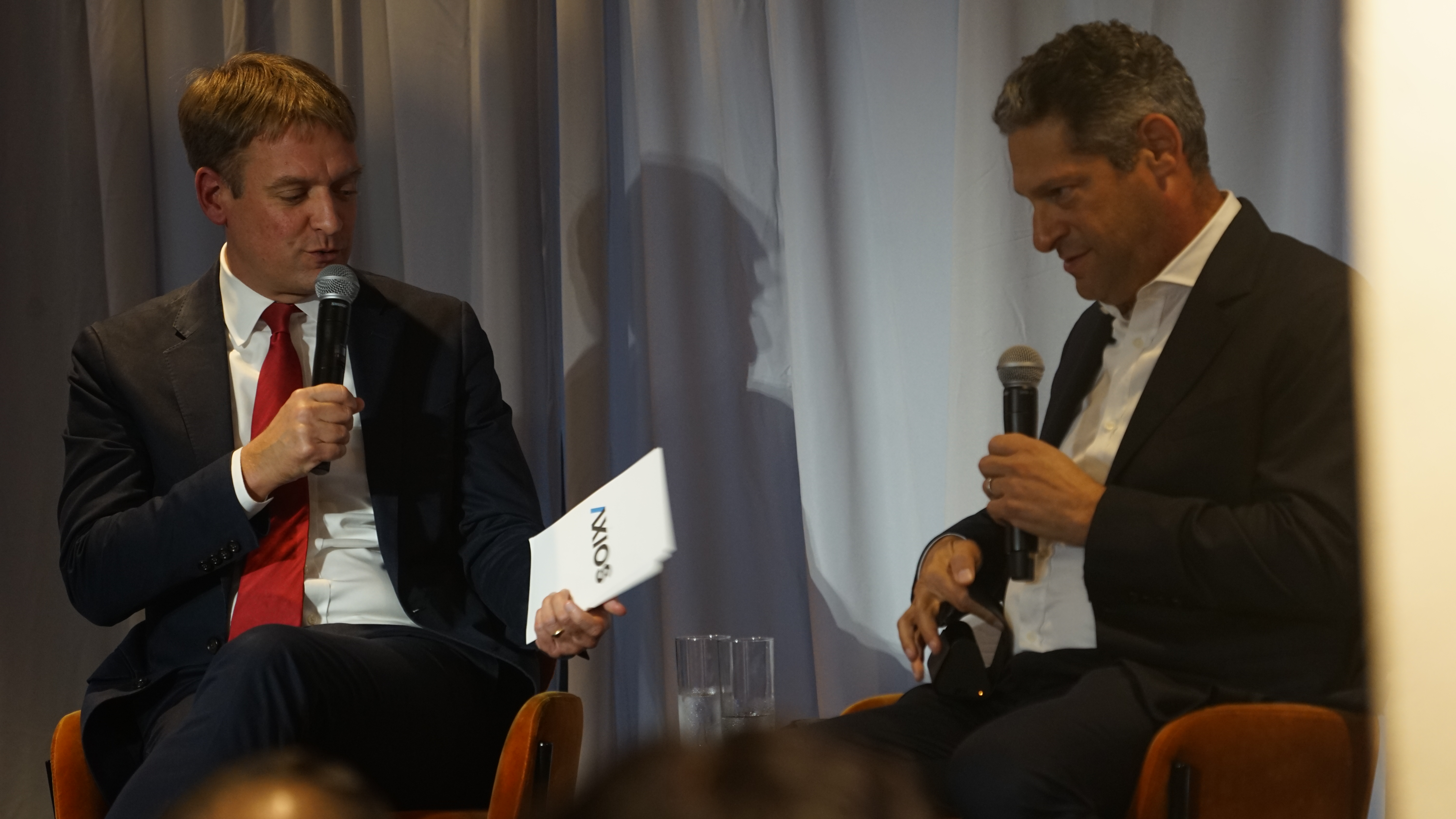 Two men in suits seated and holding microphones, speaking at an event.