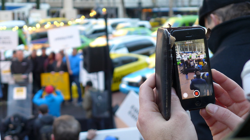 Close-up of hands holding a smartphone recording a protest with taxi cabs and people holding signs in the background.