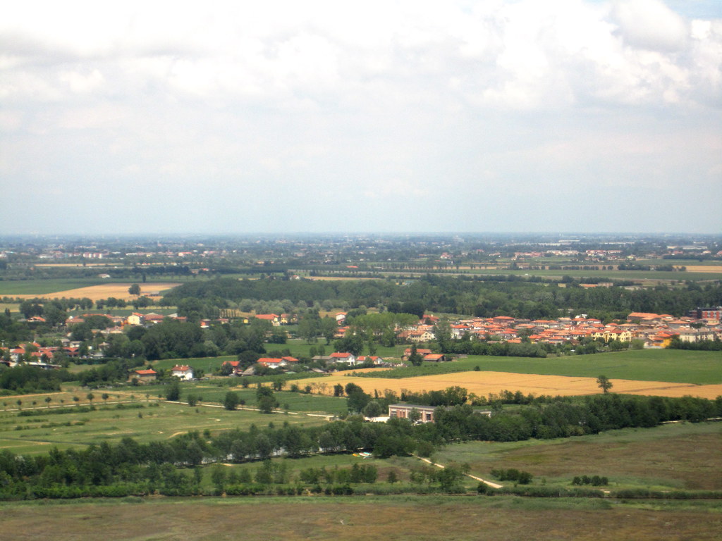 Aerial view of a small Italian village with red-roofed houses surrounded by green fields and trees under a cloudy sky.