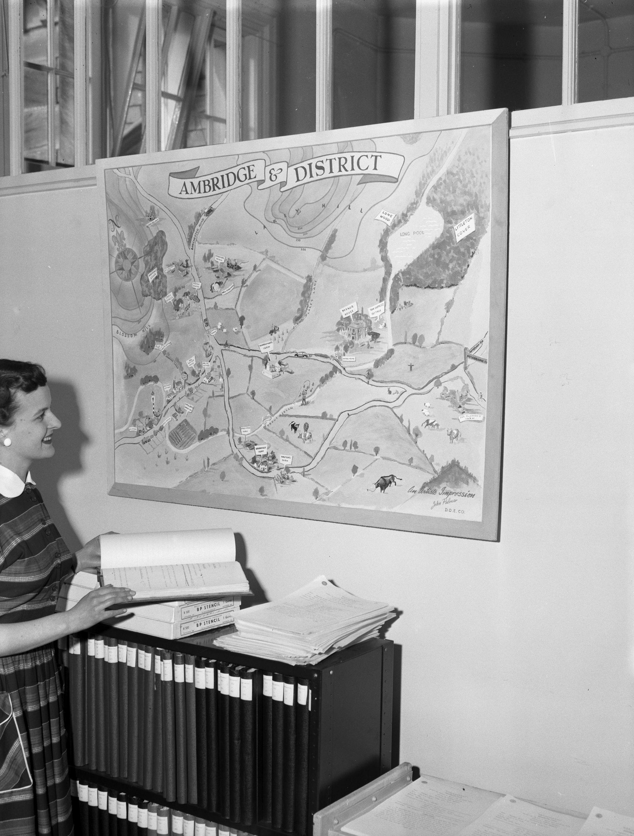 Woman looking at a large map on a wall titled 'Ambridge & District' with files and papers on a shelf beneath.