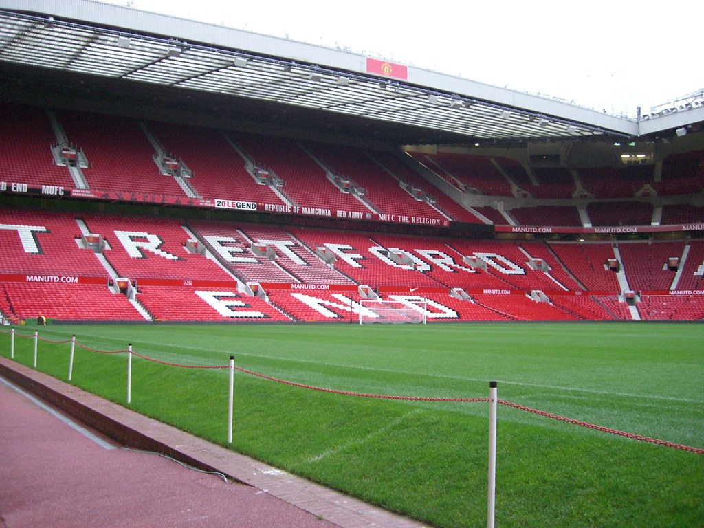 Interior view of Old Trafford football stadium showing red seats with 'OLD TRAFFORD' spelled out and the scoreboard area.