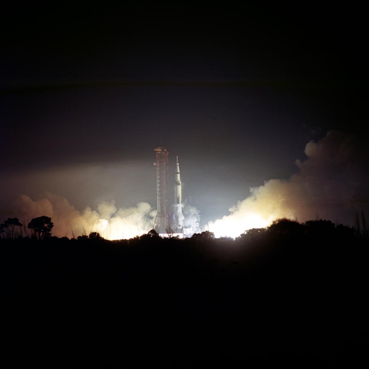 Apollo 17 rocket launching at night with smoke and bright flames at the base, launch tower visible