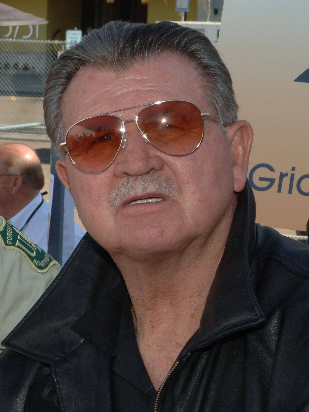 Close-up photo of an older man wearing sunglasses and a black jacket at an outdoor event.