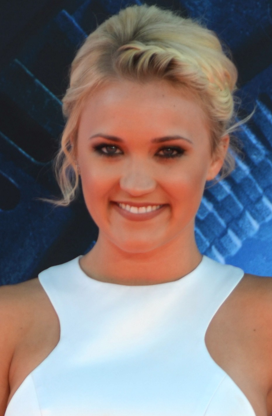 Headshot of a smiling woman with blonde hair in an updo wearing a white sleeveless dress against a dark blurred background.