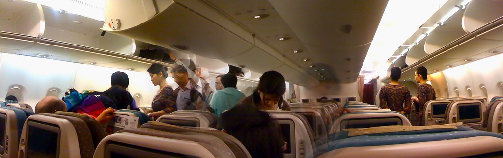 Panoramic view of an airplane interior with passengers and crew, showing seats and overhead compartments.