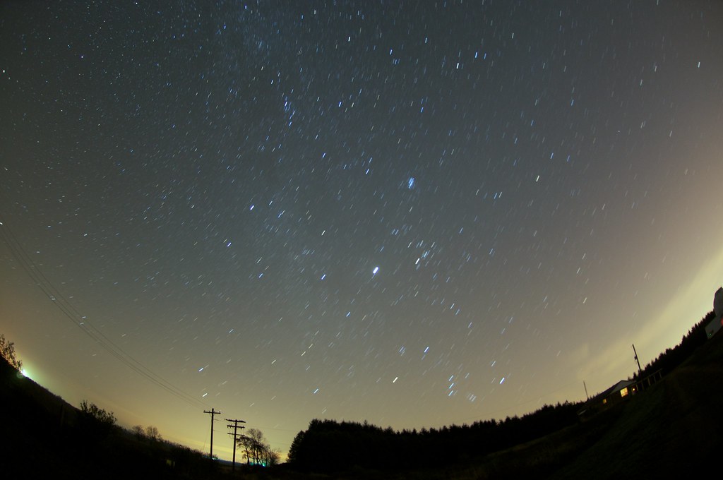 Night sky with star trails over a rural landscape with power lines and trees