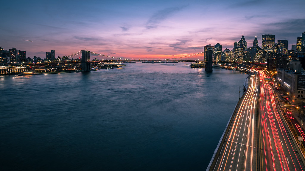 Sunset view of Manhattan skyline and Brooklyn Bridge with river and traffic light trails.