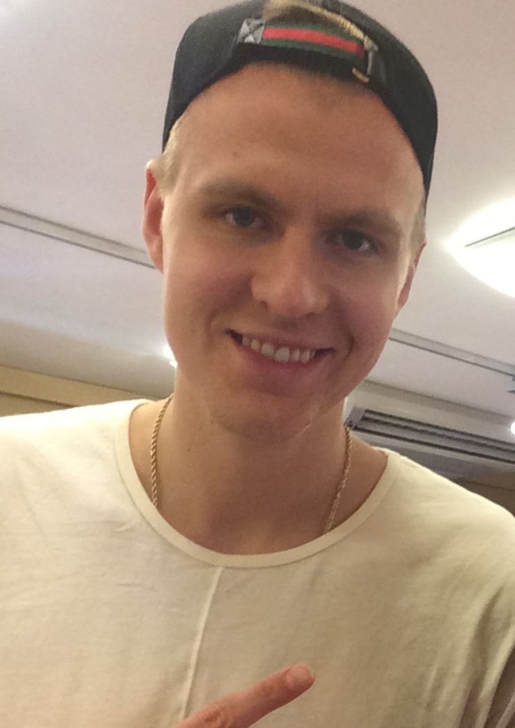 Close-up photo of a smiling young man with a backward black cap, white shirt, and a gold chain, pointing with his finger.