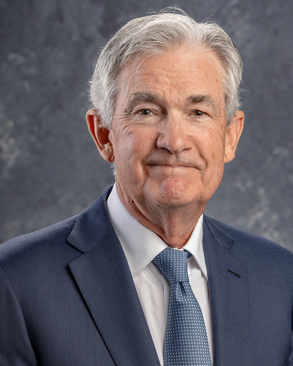 Head and shoulders portrait of an older man with gray hair, wearing a blue suit, white shirt, and blue patterned tie, against a mottled gray background
