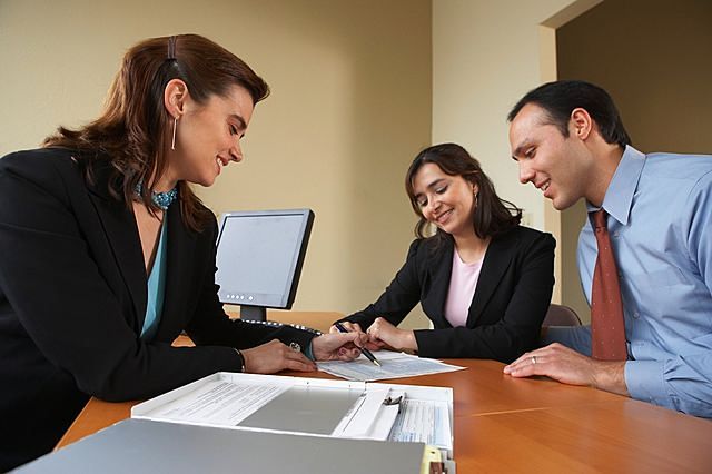 A couple sitting at a desk with a real estate agent who is pointing at documents.
