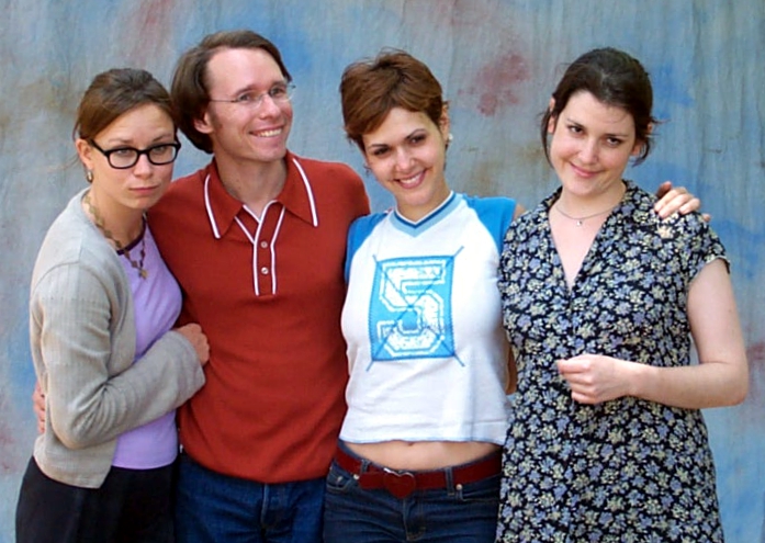 Four people standing close together, three women and one man, smiling in front of a blue-gray backdrop.
