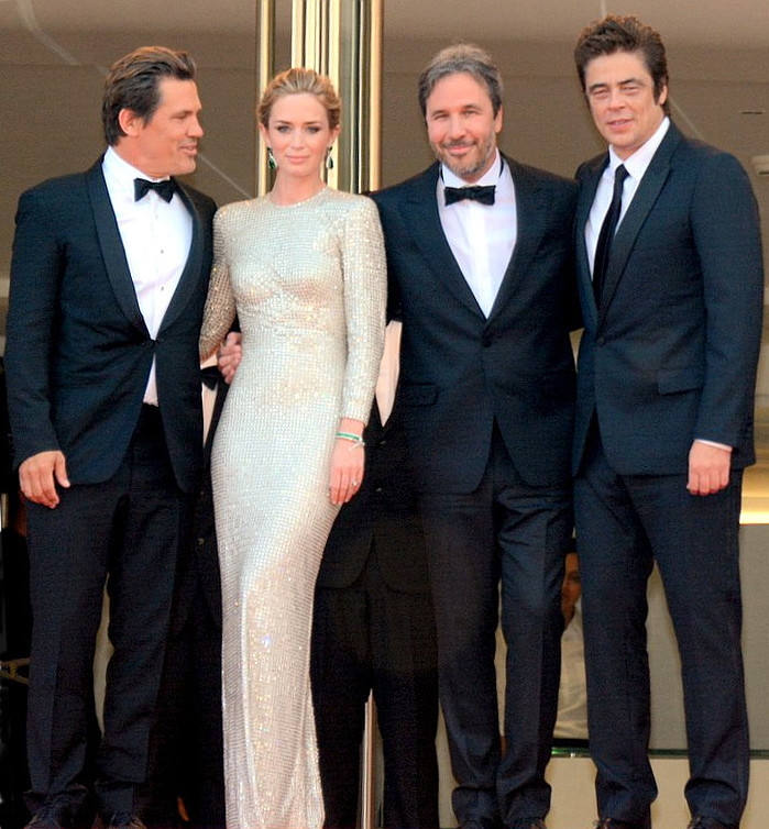 Four people in formal attire posing at a venue entrance. Two men in tuxedos flank a woman in a shiny, long-sleeved gown, and another man in a tuxedo stands on the far right.