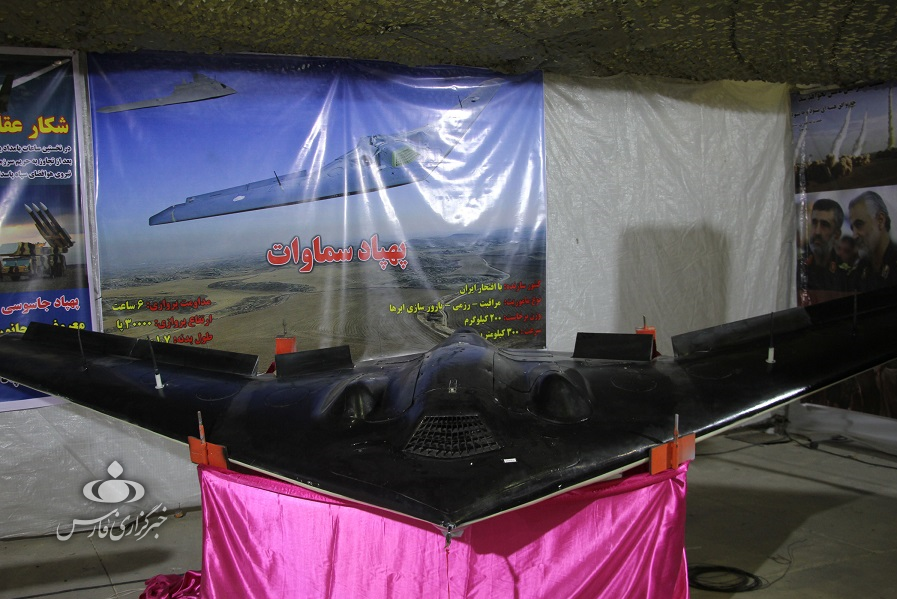 A black Shahed Samavat UAV drone displayed indoors on a pink-covered table, with a large poster behind it showing the drone flying over a landscape.