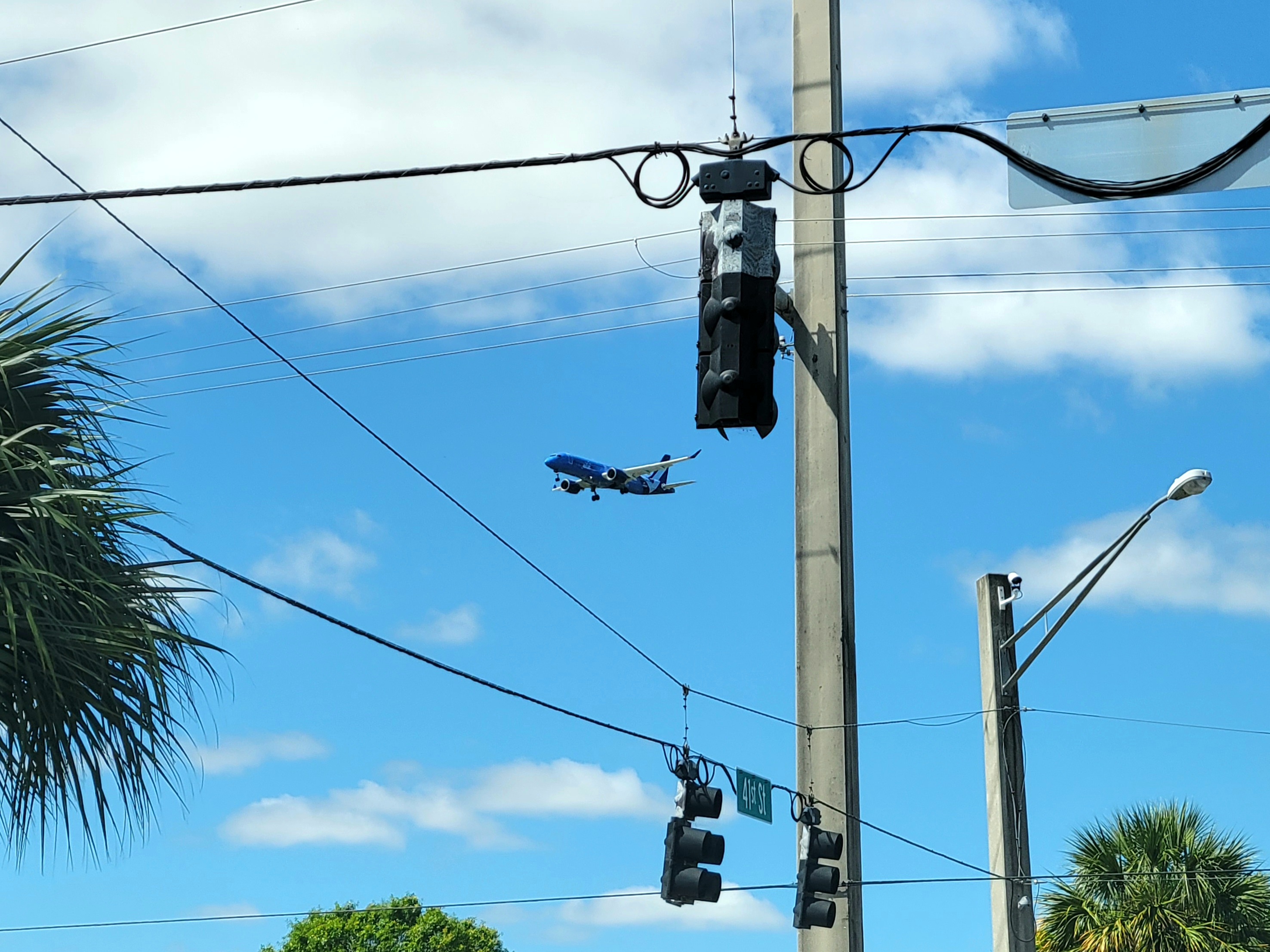 A blue Breeze Airways airplane flying in the sky with traffic lights and palm trees in the foreground.