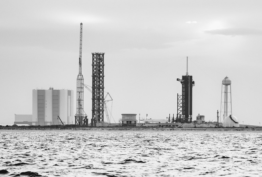 Black and white photograph of SpaceX Starship launch tower construction at Launch Complex 39A, with water in the foreground.