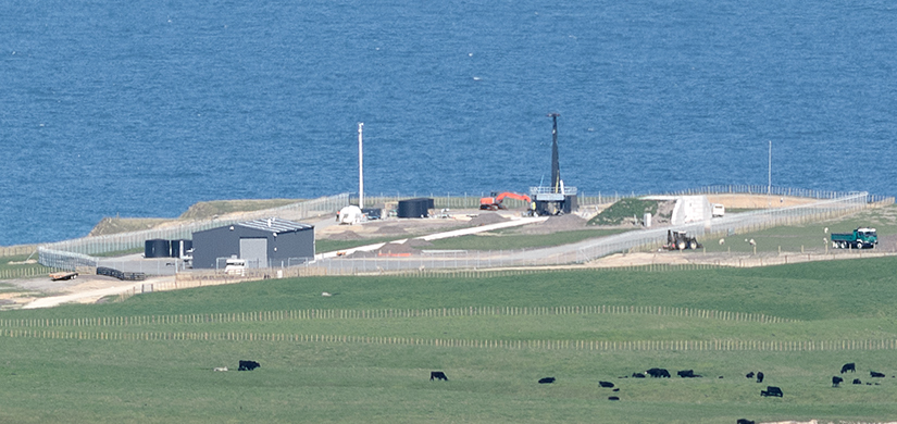 A fenced launch site with buildings, a tall black launch platform, located on green grass near the ocean, with grazing cattle nearby.