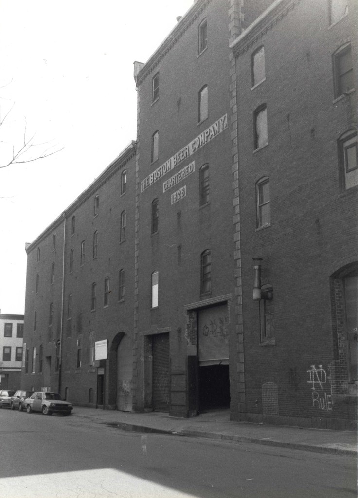 Black and white photo of a multi-story brick building with a sign reading 'The Boston Beer Company Chartered 1828' on its facade, located along a street with parked cars.