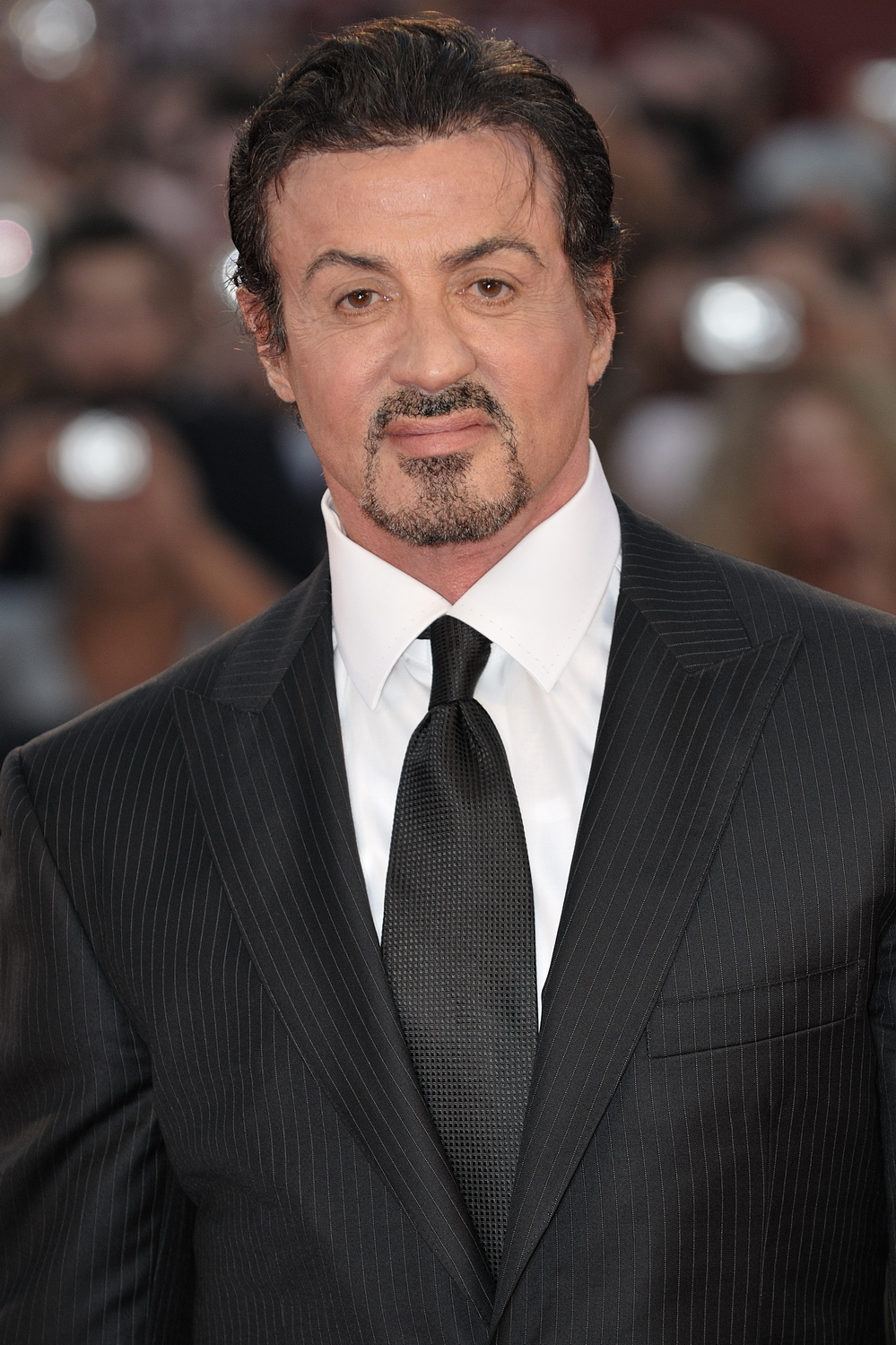 Close-up portrait of a man with dark hair and a goatee, wearing a black pinstripe suit, white shirt, and black tie, at a red carpet event.