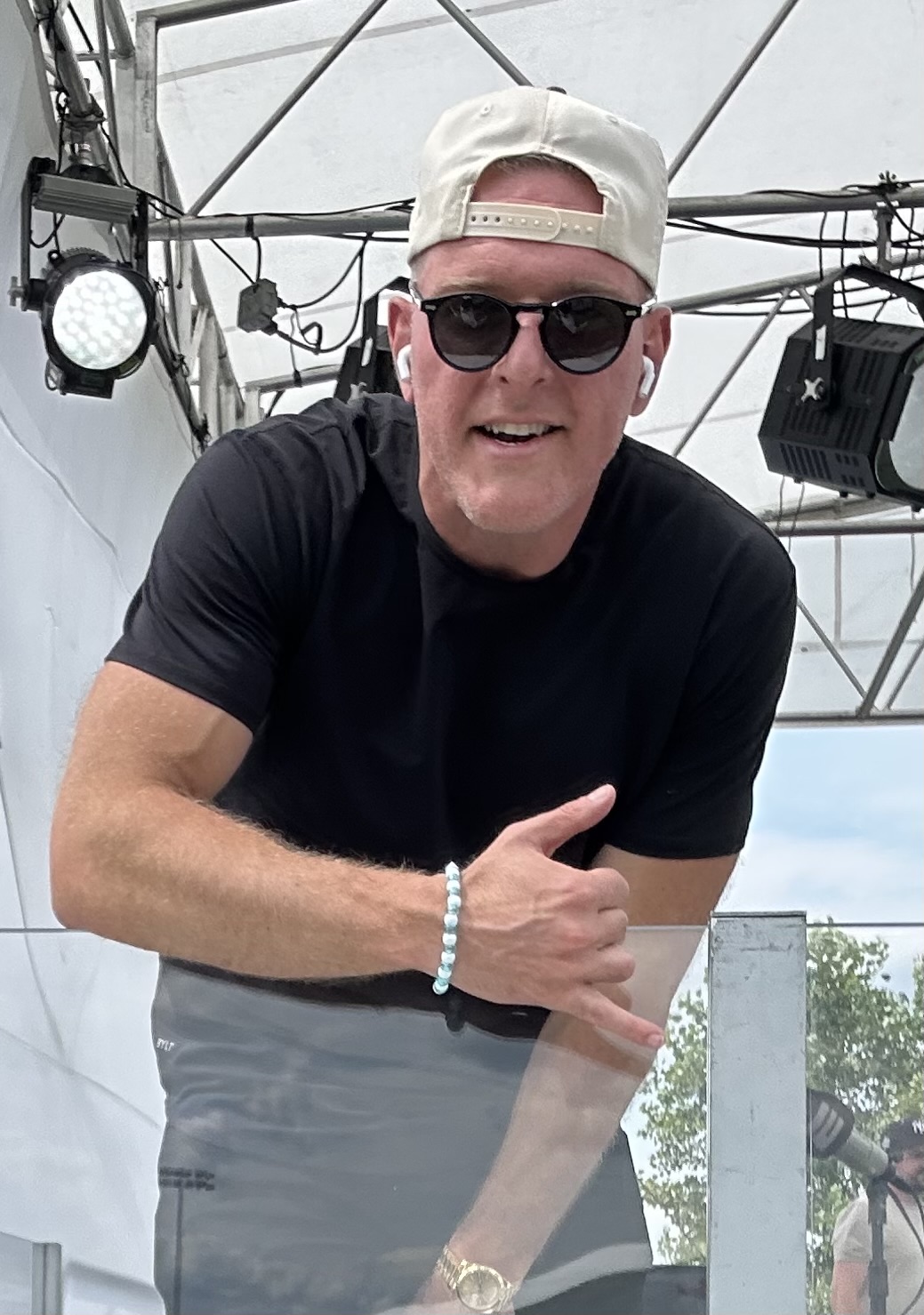 Man wearing a backward beige cap, black sunglasses, a black t-shirt, and a bracelet, leaning forward with a hand gesture under a tent structure.
