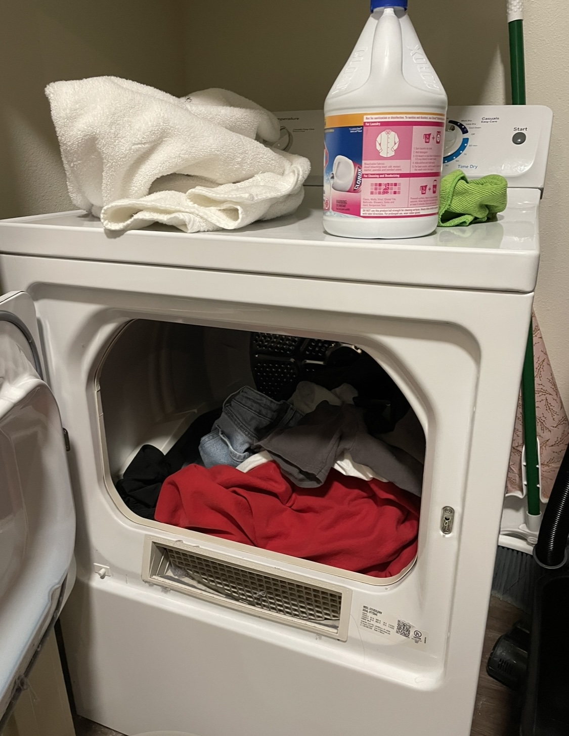 Open clothes dryer with various clothes inside, white towels and a bottle of cleaner on top.