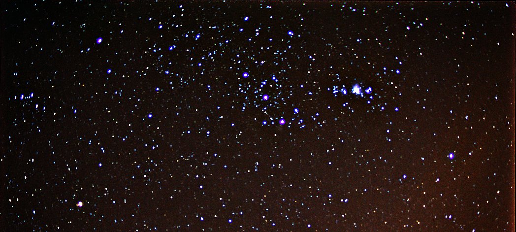 Night sky photograph showing the Orion constellation with many stars visible, slightly blurry, faint orange tint at edges