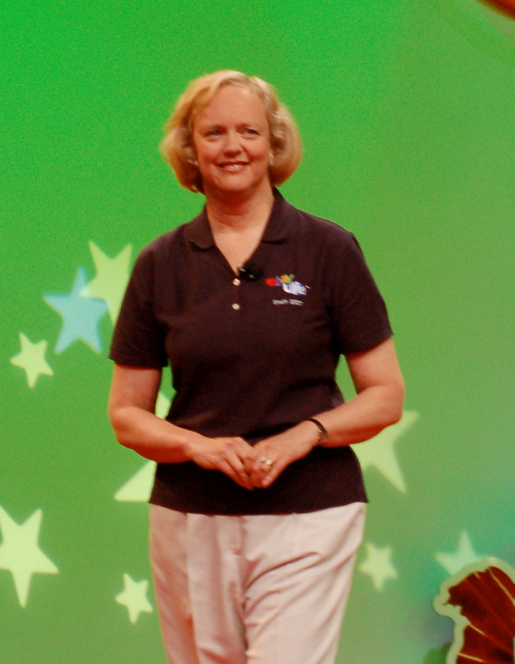 Meg Whitman standing with a green background with stars, wearing a black polo shirt and light pants.
