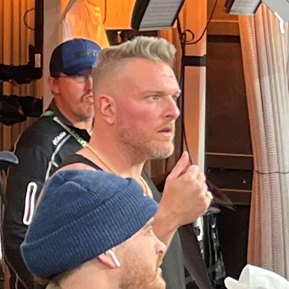 Man with blond hair and beard giving a thumbs up, standing indoors with two other men in the background