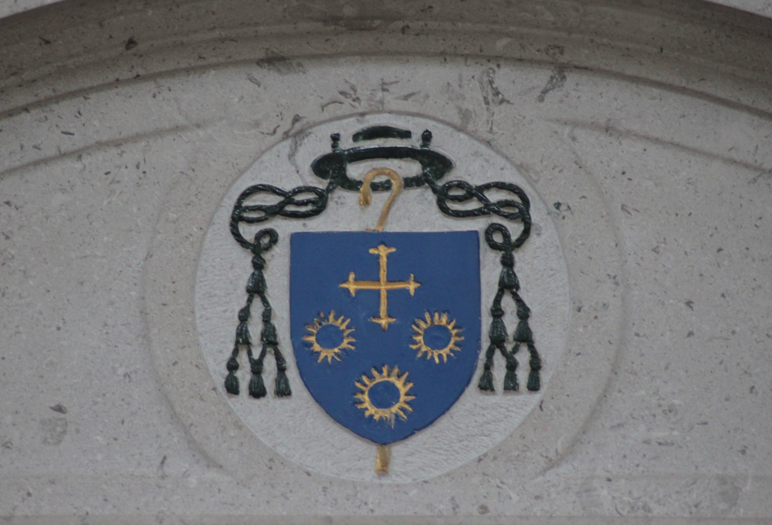 Close-up of the stone carving of the arms of the Bishop of Brentwood on the cathedra, featuring a blue shield with a gold cross, gold sun symbols, and a hat with tassels.