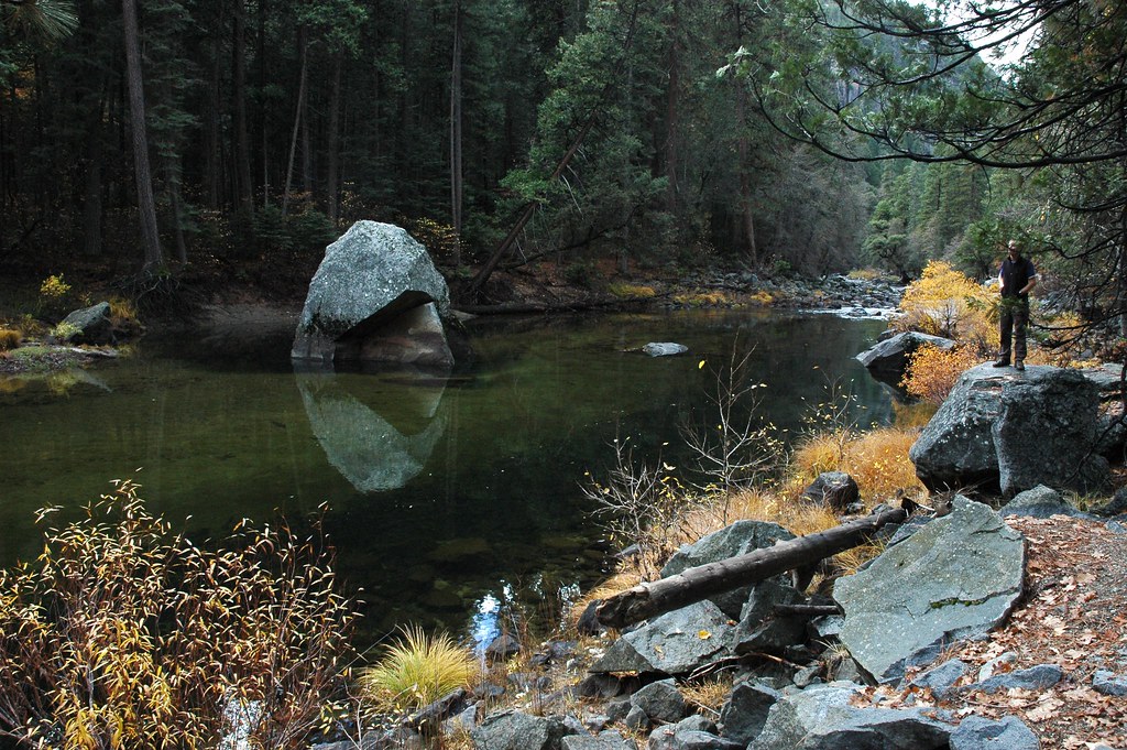 A visitor standing on a rock beside a calm river with large granite stones and fall colors in Yosemite National Park.