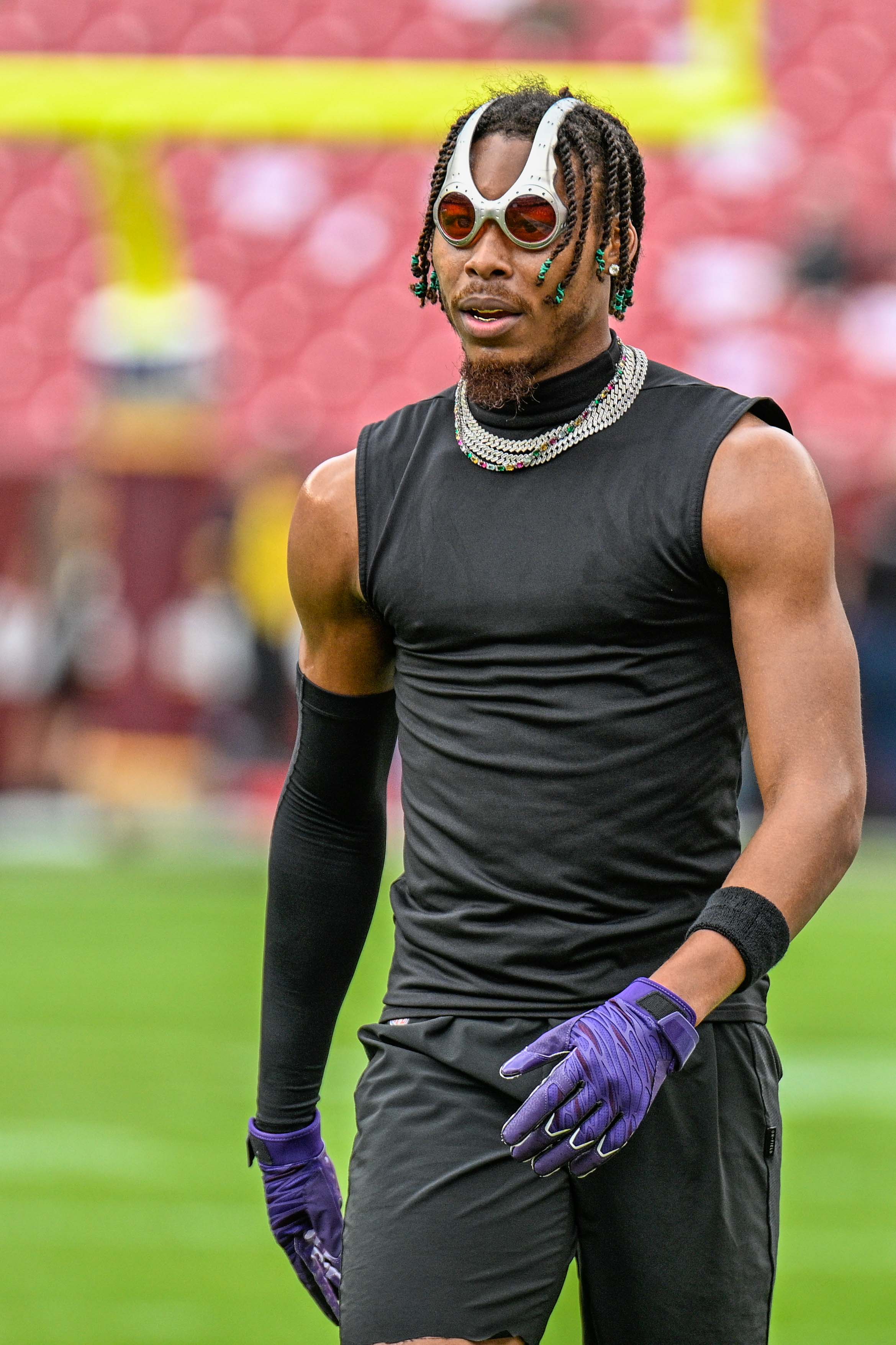 A man in a sleeveless black shirt, purple gloves, and unique sunglasses stands on a football field with a blurred stadium background.